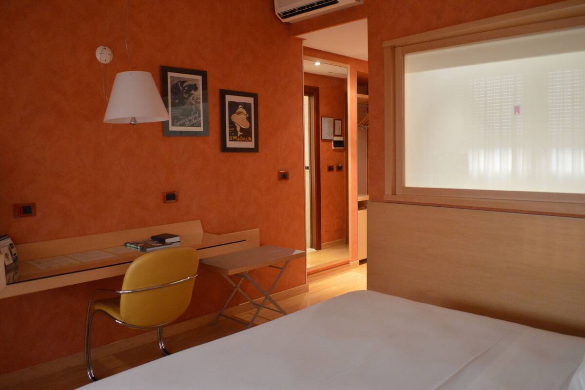 Una habitación de hotel con una pared naranja, una cama, un escritorio con una silla, una ventana con persianas y una puerta que da a otra habitación.