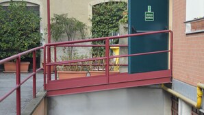 Una escalera estrecha conduce a una puerta de garaje, con una barandilla roja a la izquierda y un letrero de salida de emergencia verde arriba.
