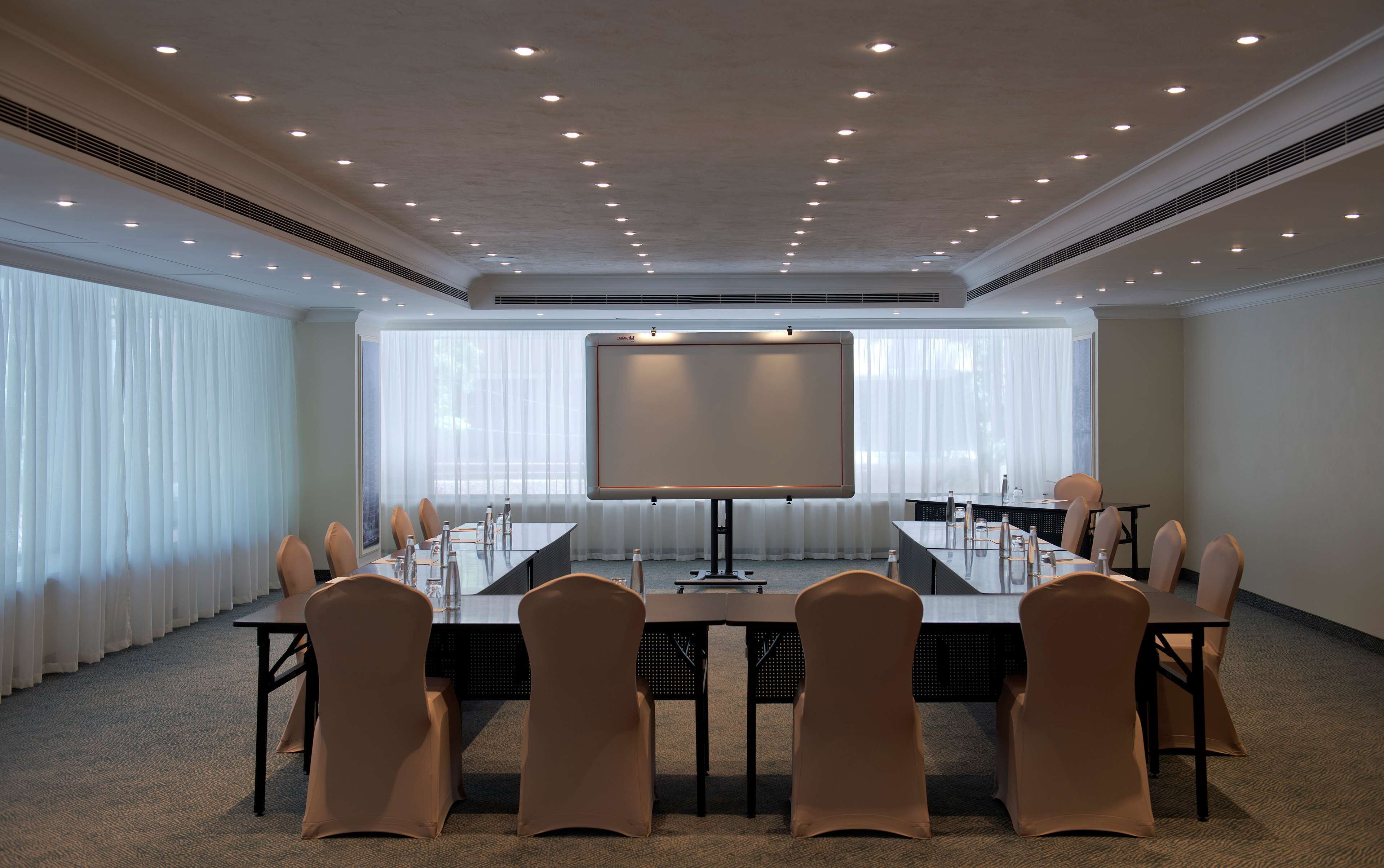 Un salón de conferencias con una larga mesa, sillas, una pantalla de proyector y un amplio ventanal con cortinas.