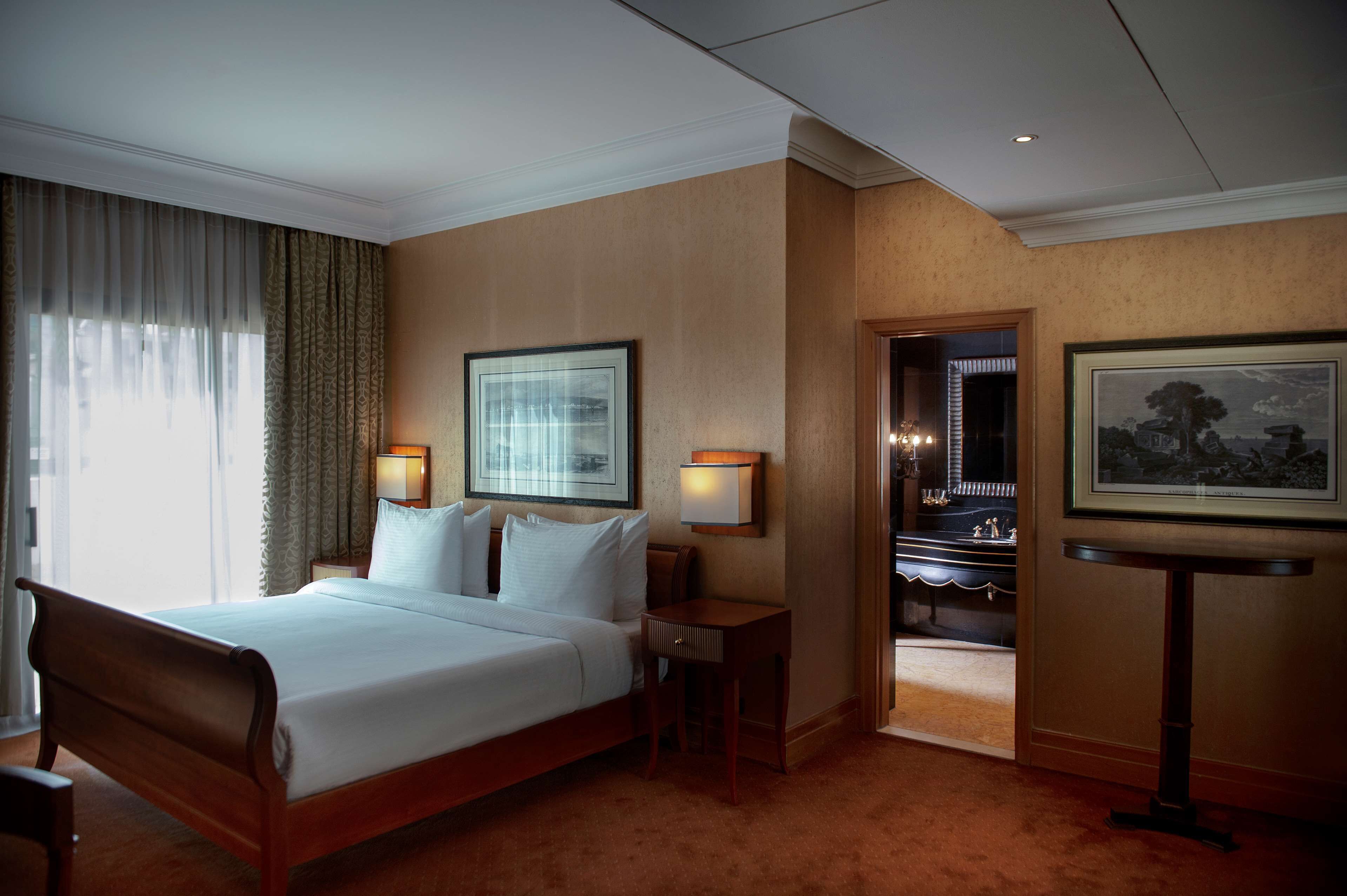 Een hotelkamer met een groot bed, twee nachtkastjes, een nachtkast en een inlijsting afbeelding aan de muur, plus een deur naar een badkamer.