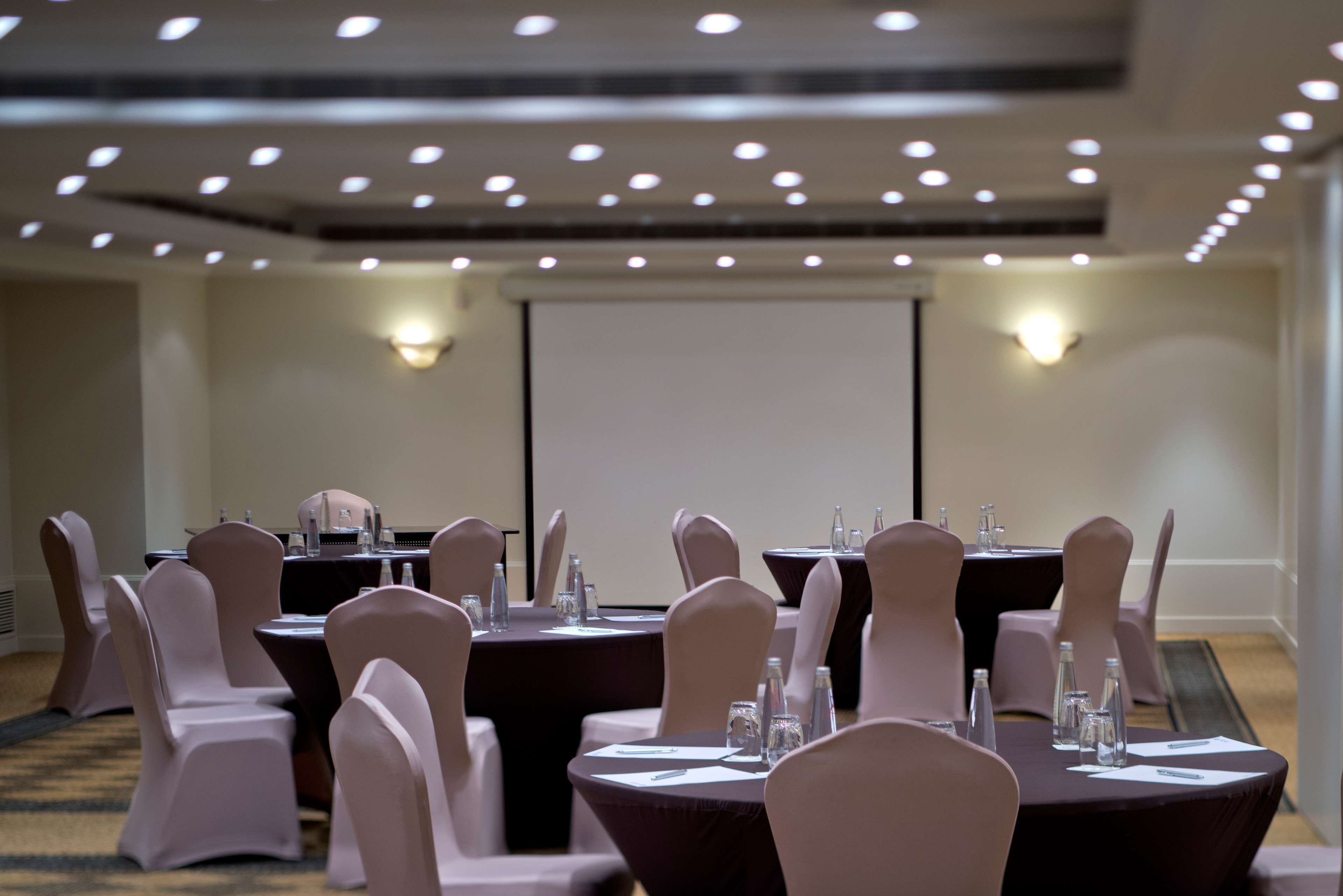 Un salón de conferencias con mesas redondas, sillas y una pantalla para proyector.