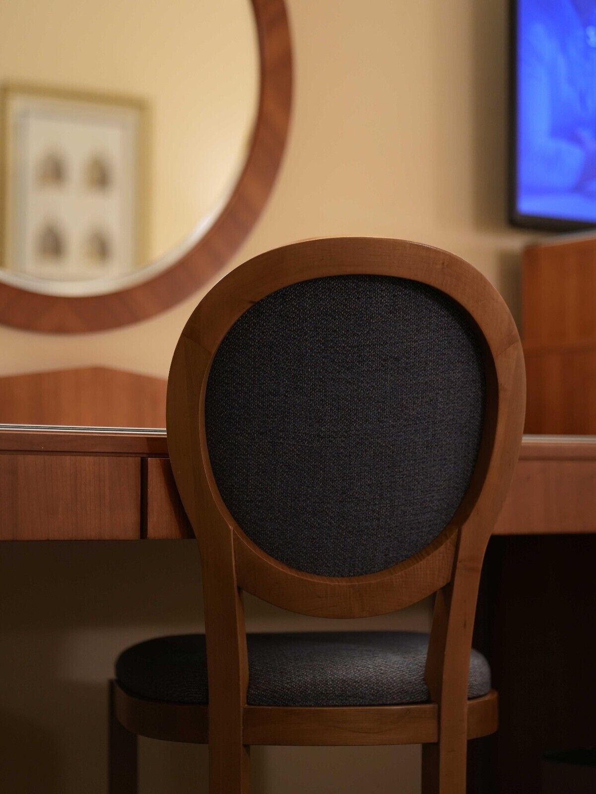 A wooden chair with a dark fabric backrest and seat, positioned in front of a desk with a round mirror on the wall.