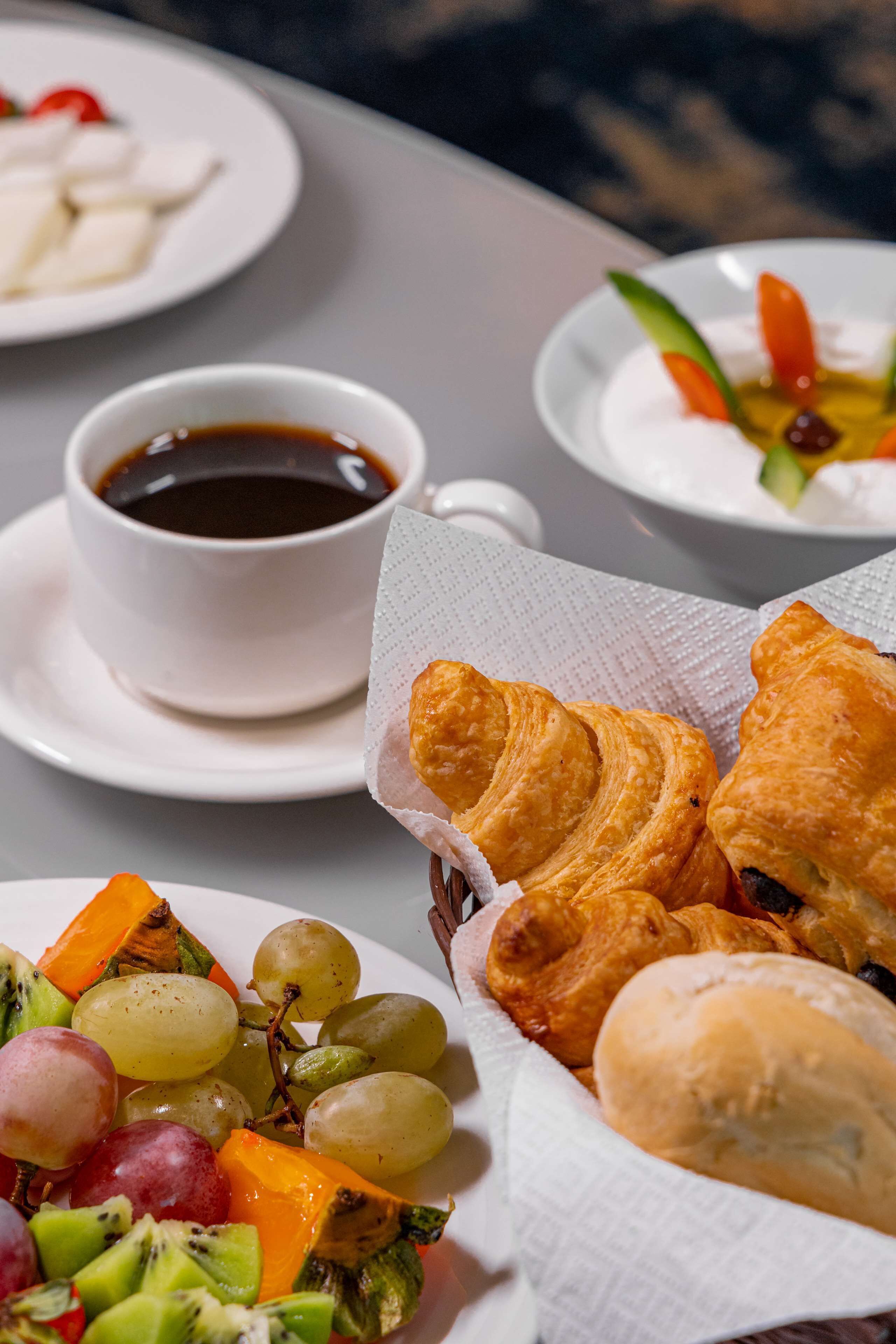A table with plates of food, including croissants, grapes, and a salad, and a cup of coffee.