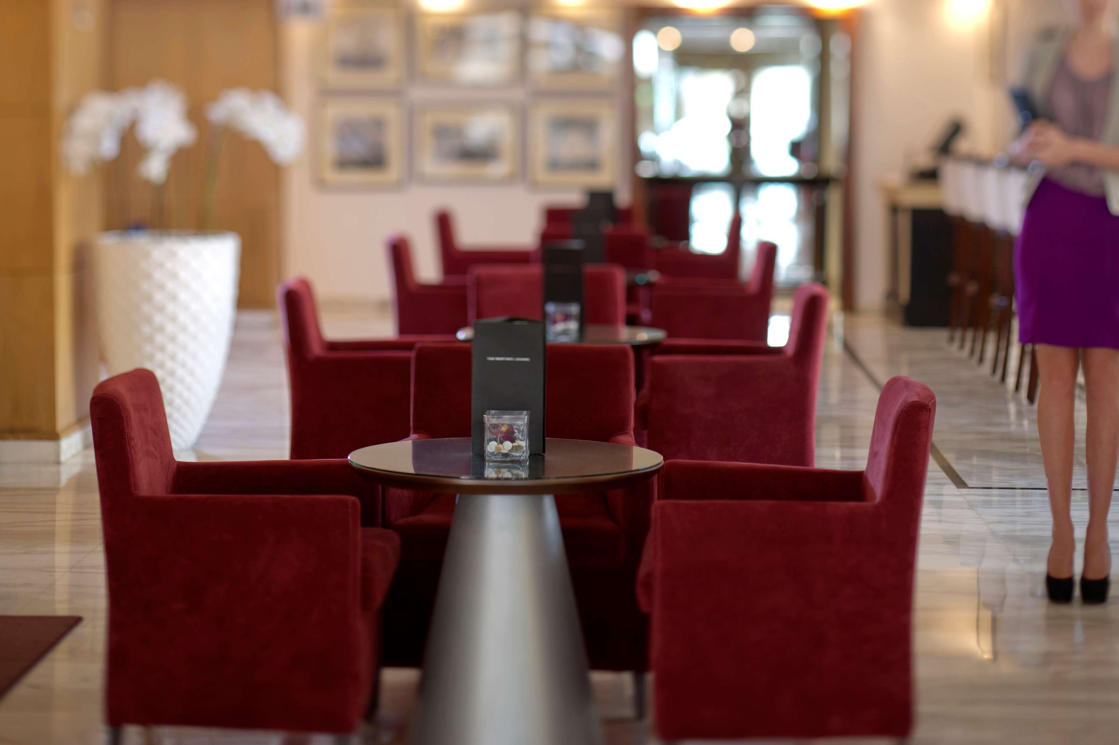 A hotel lobby with red upholstered chairs arranged around a central table, a reception desk, and framed pictures on the wall.