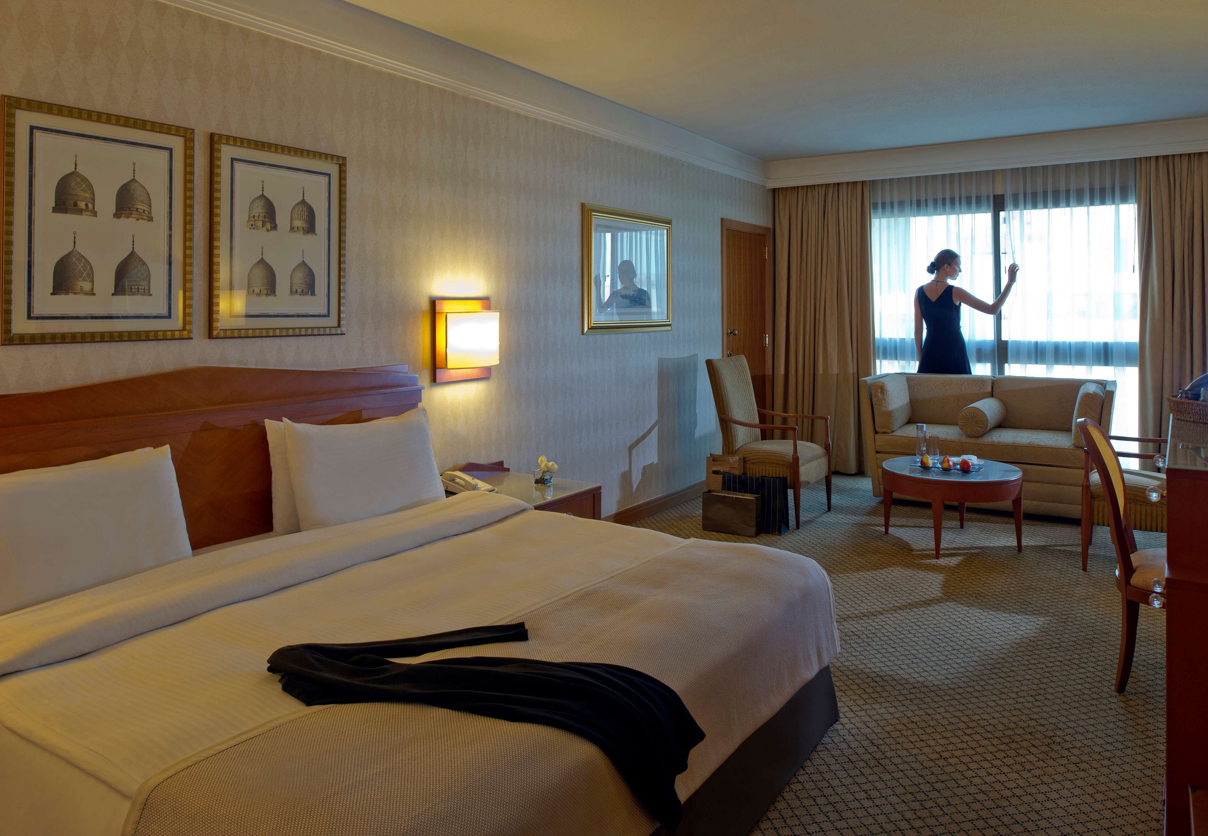 Habitación de hotel con una cama grande, una zona de estar con sofá y sillas, una mesita y cuadros en las paredes.