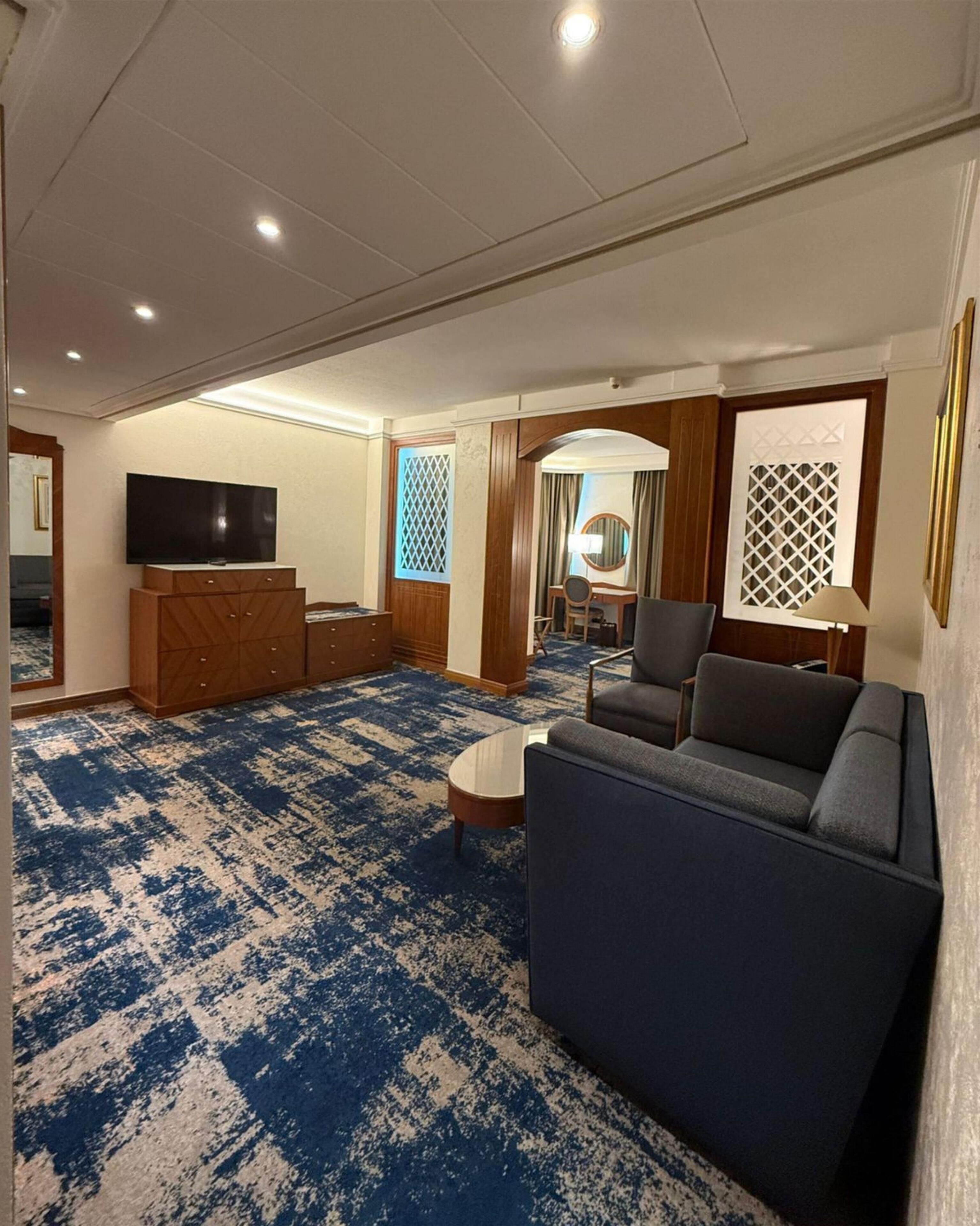 A hotel room with a large blue patterned carpet, a sofa, a television, a desk, and a chair.