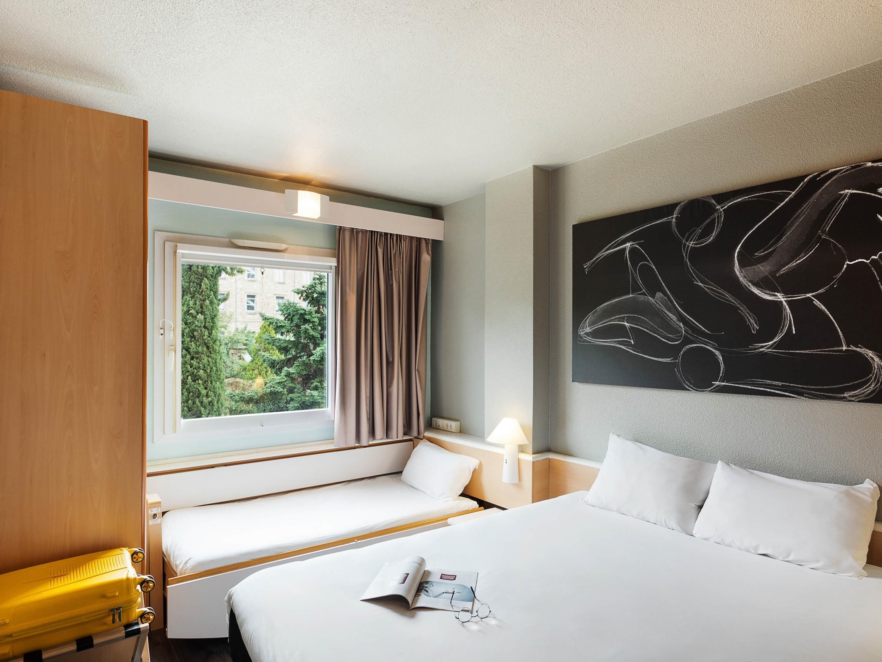 Une chambre d’hôtel avec un lit, une fenêtre avec des rideaux, une table de chevet avec une lampe et une œuvre d’art décorative au mur.