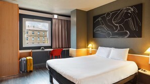 Une chambre d’hôtel avec un grand lit, un bureau avec une chaise, une fenêtre avec des rideaux, une œuvre d’art accrochée au mur et une table de chevet avec une lampe.