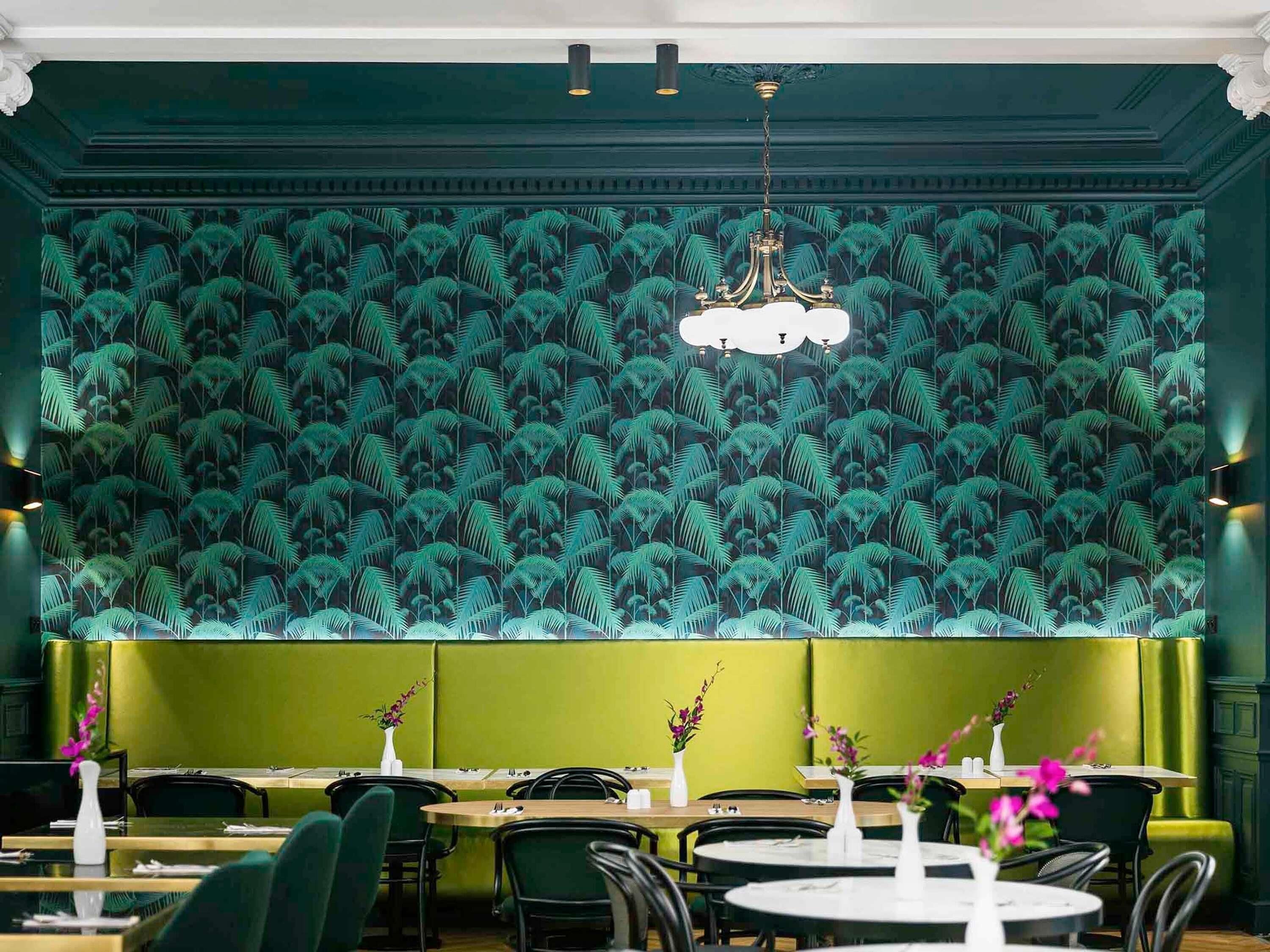 Een restaurant met groene wanden, behang met patroon en een kroonluchter. Tafels zijn gedekt met witte vazen en bloemen.