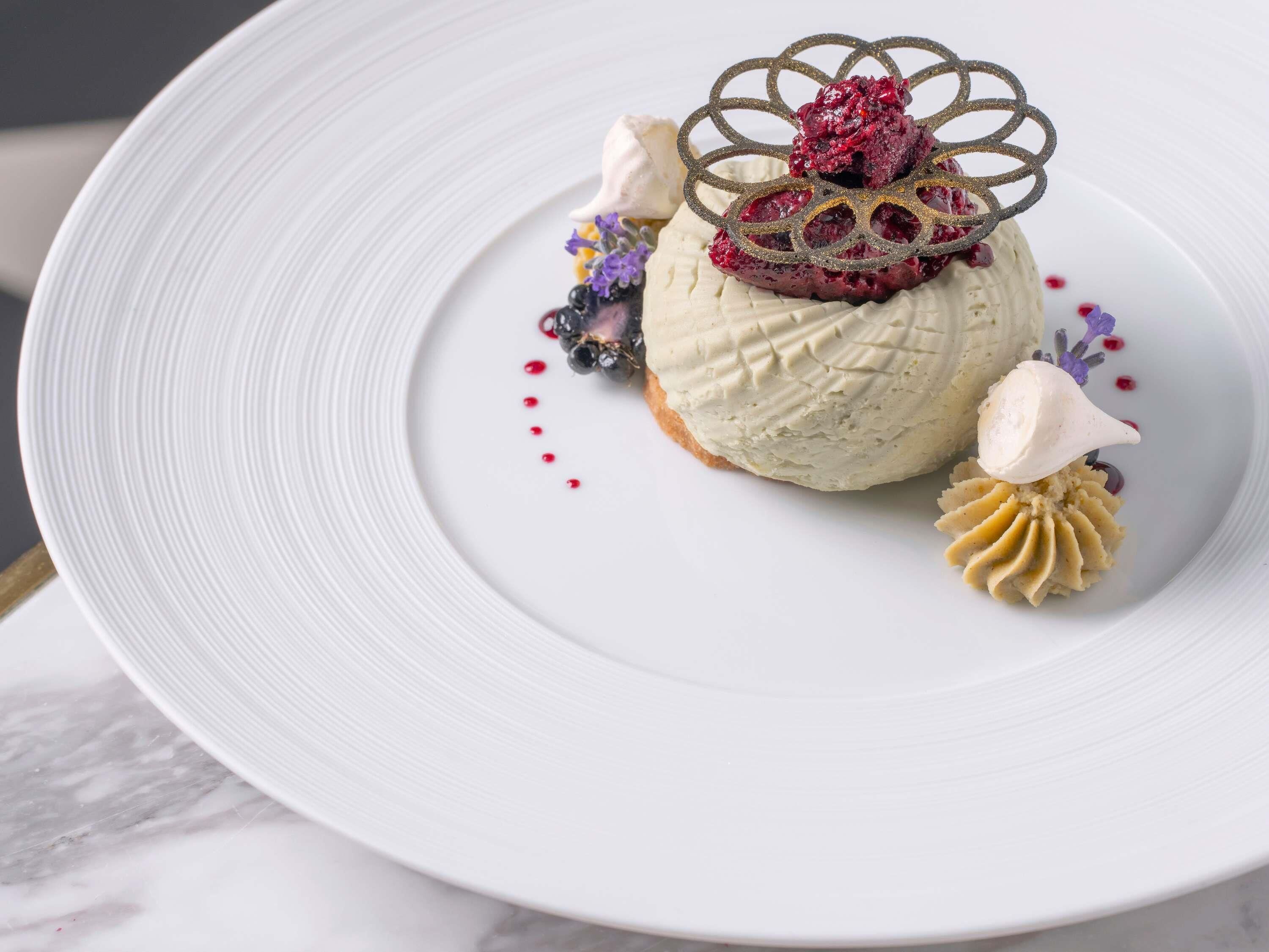 Ein Dessert auf einem weißen Teller mit einem verzierten Metallteil, Beeren und einem Klecks Sahne.
