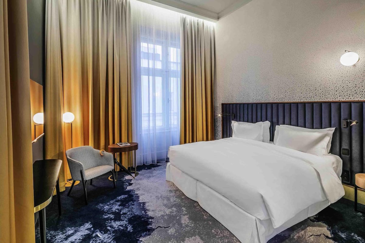Quarto de hotel com uma cama grande, uma cadeira, uma mesa pequena e uma janela com cortinas.
