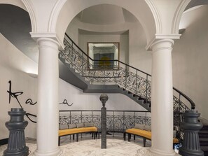 A grand staircase with ornate black railings, a marble floor, and a bench area with a yellow cushion. - Century Old Town Prague - MGallery Collection (Prague)
