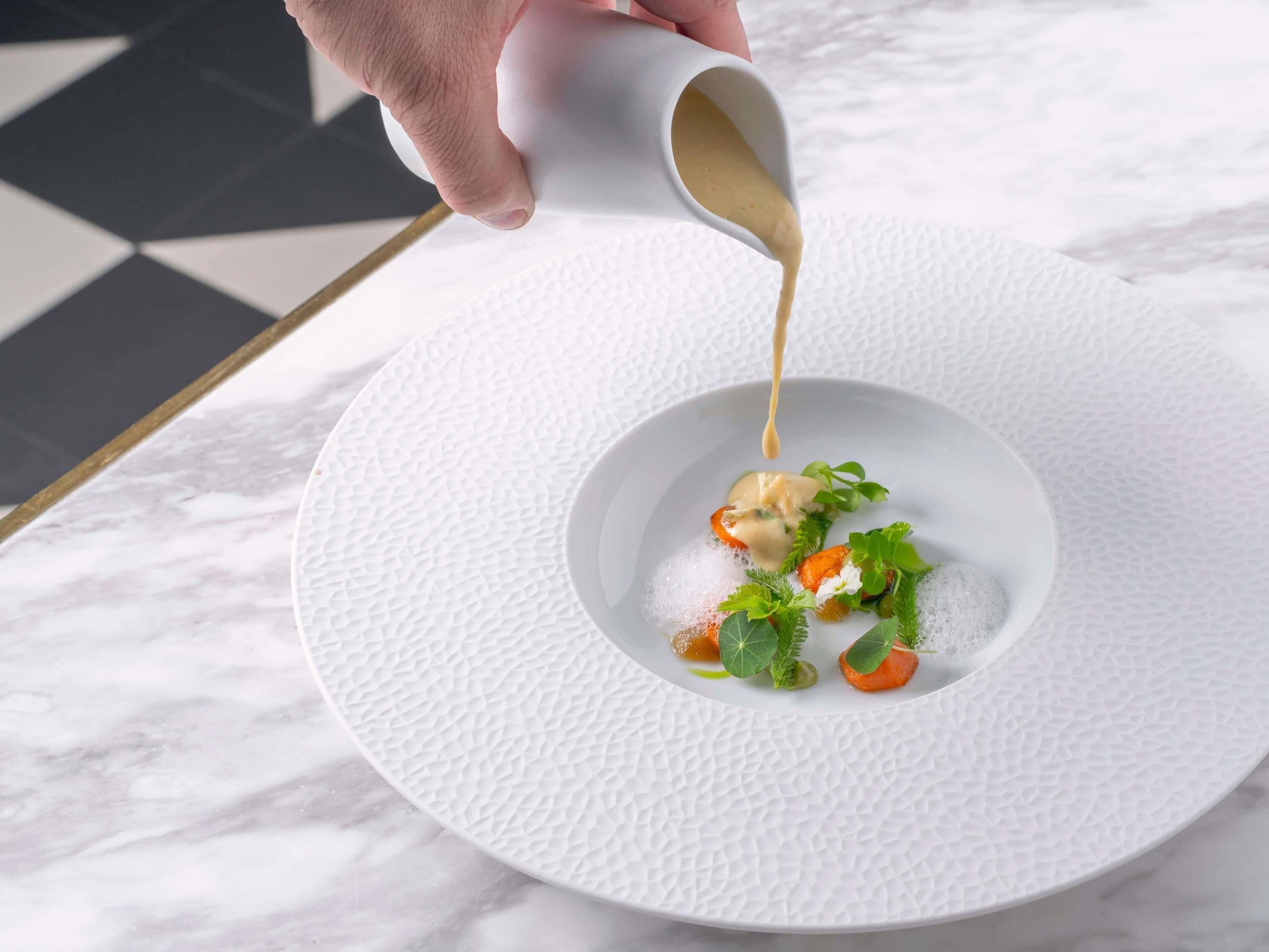 Een witte, gestructureerde schaal met een kleine kom, gevuld met eten, versierd met groenten, en overgoten met saus.