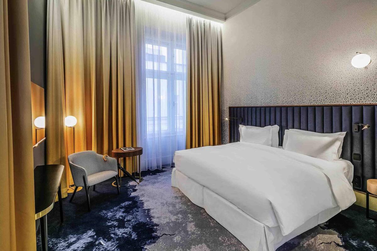 Habitación de hotel con una cama grande, una silla, una mesita y una ventana con cortinas.