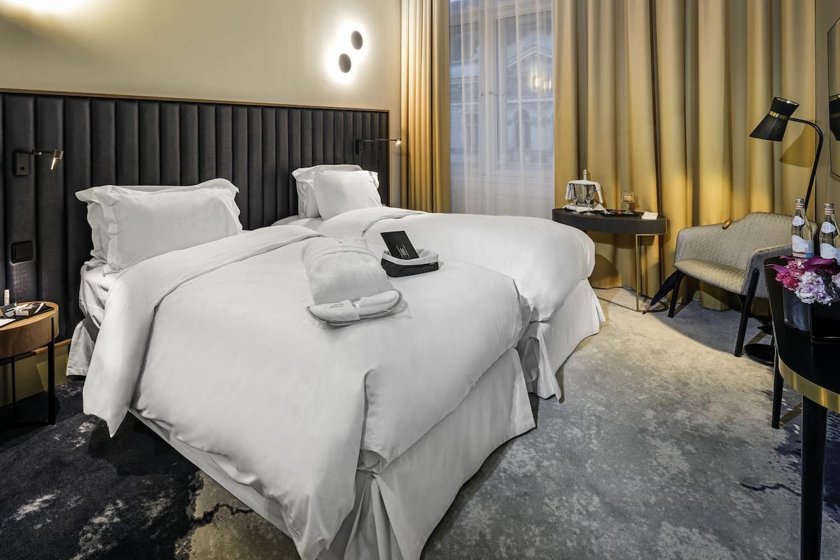 Habitación de hotel con una cama grande, una mesita de noche con lámpara, una silla y vista a un edificio a través de la ventana.