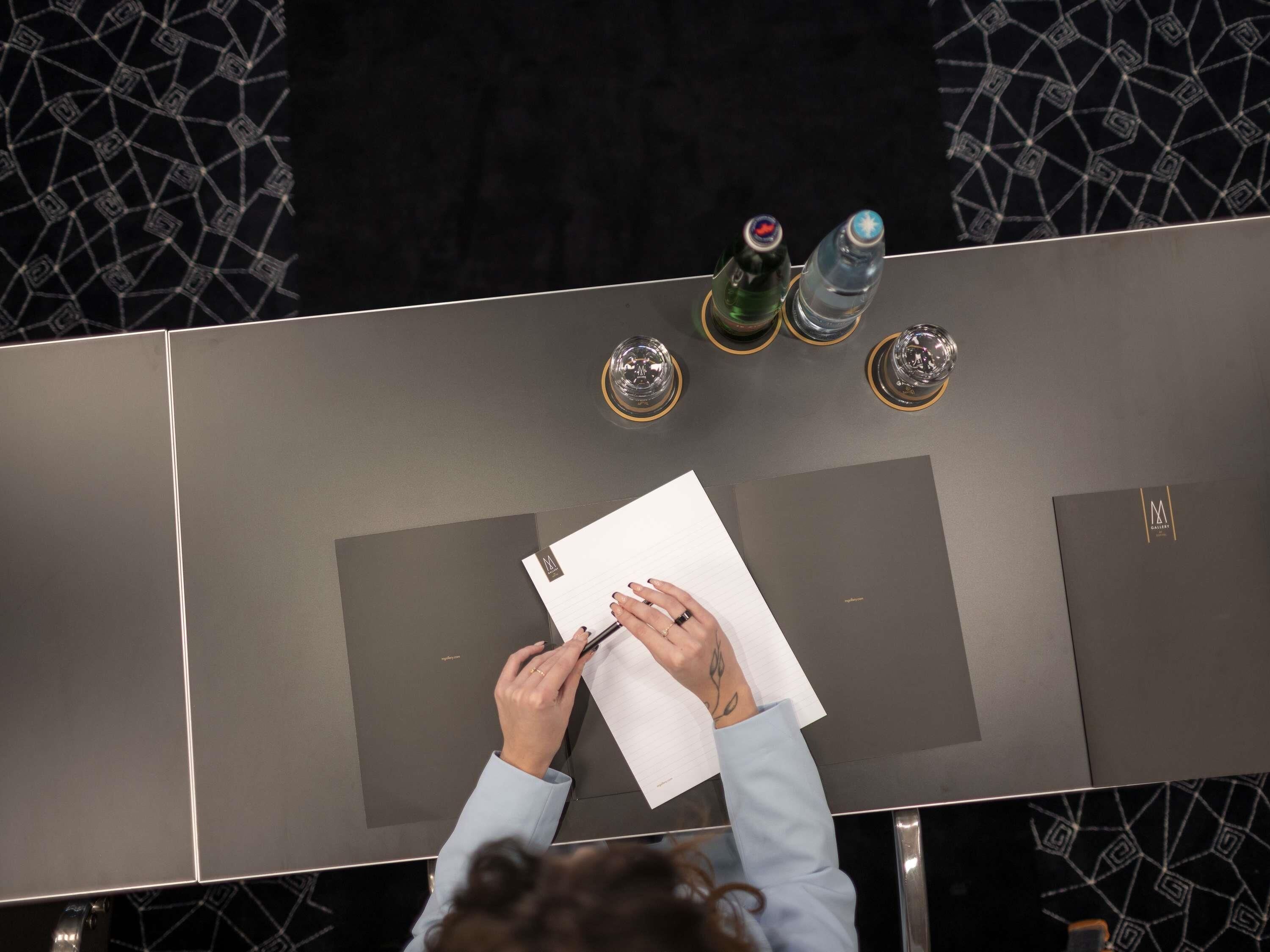 A person writing at a table with a notebook, glasses, and two bottles of water.