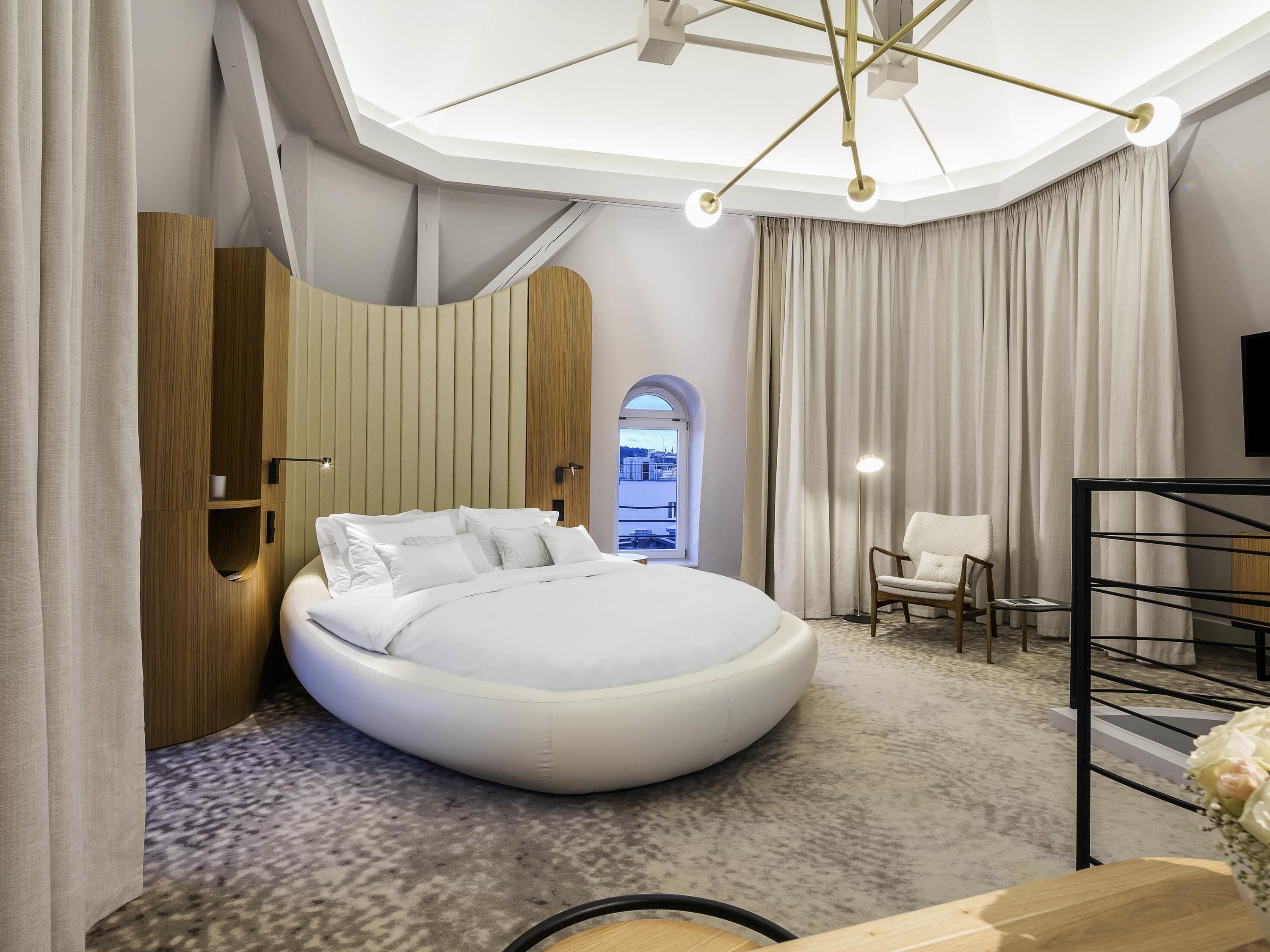 Een moderne hotelkamer met een rond bed, een houten garderobe, een zithoek met een fauteuil en een uitzicht door een gebogen raam.