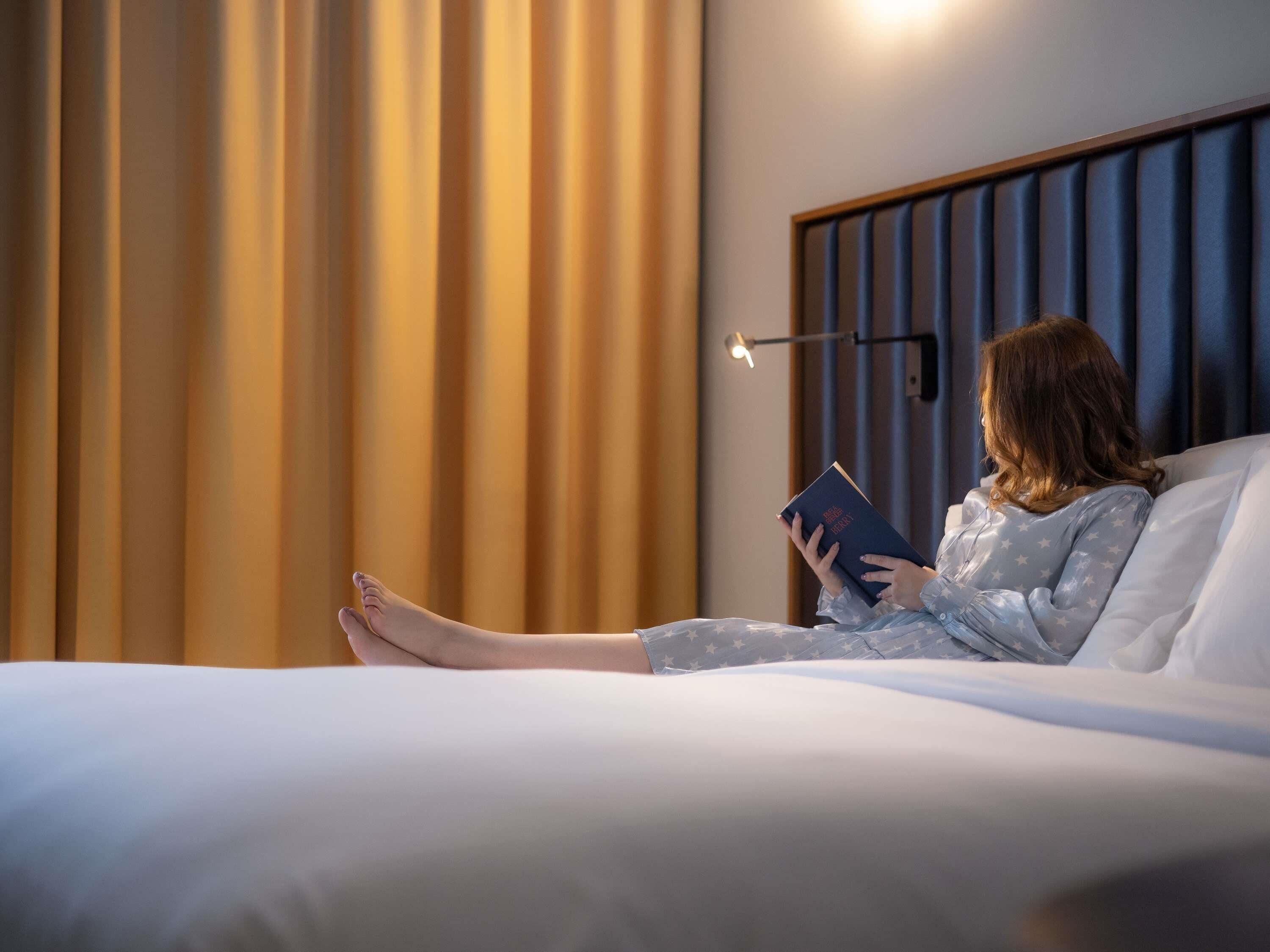 A person lying on a bed in a room with a headboard, reading a book.