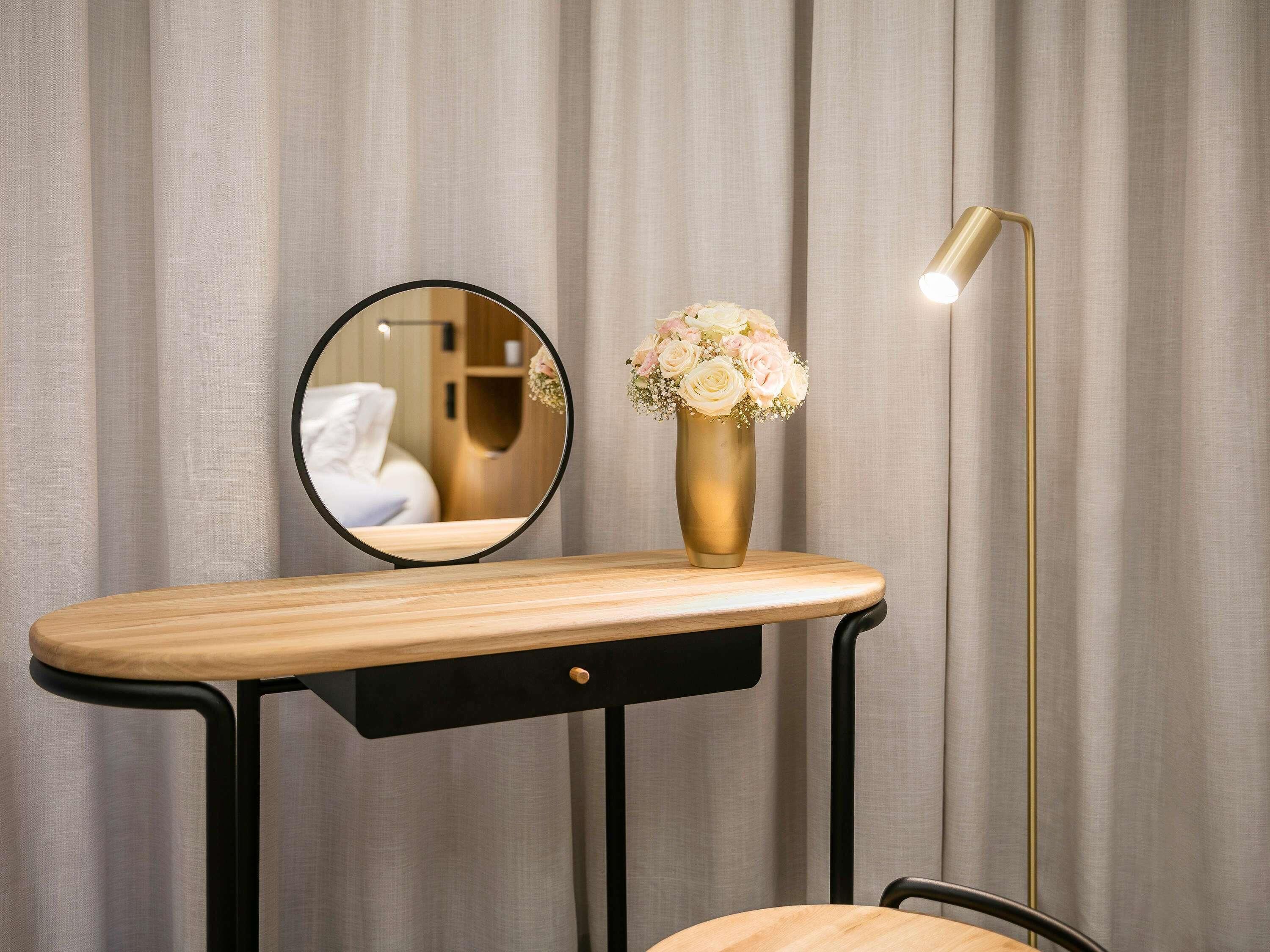 Une coiffeuse avec un miroir, un vase de fleurs et un lampadaire, le tout placé devant un rideau.