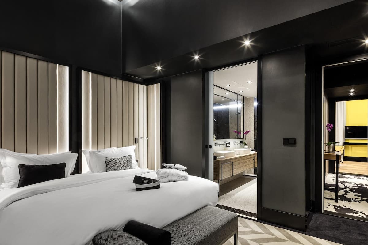Habitación de hotel moderna con una cama grande, mesitas de noche, un baño con espejo y vistas al pasillo.