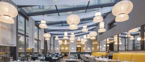 A modern restaurant with a high ceiling, glass panels, and multiple hanging light fixtures. Tables are set with white tablecloths and chairs.
