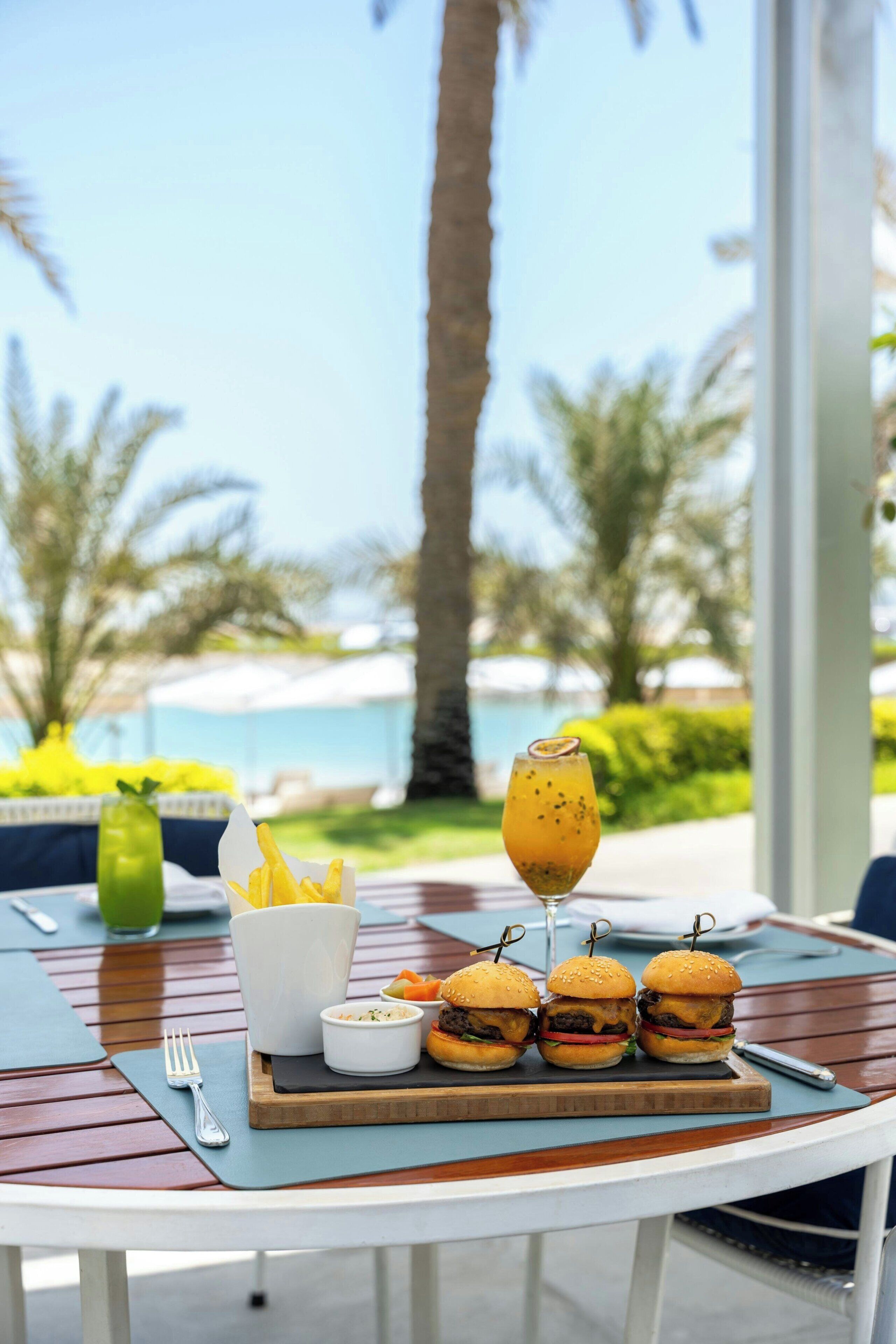 A table with a meal consisting of burgers, fries, and a drink, set against a backdrop of a pool and palm trees.