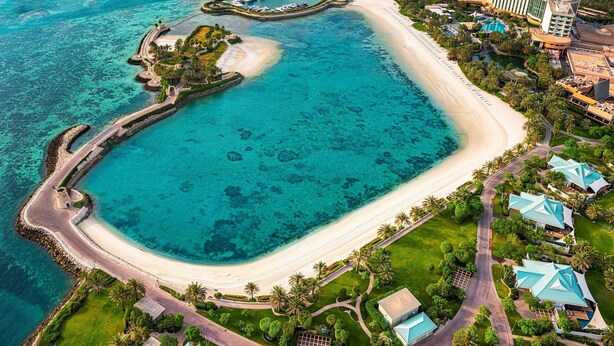 An aerial view of a coastal resort with a sandy beach, clear turquoise waters, and a well-maintained landscaped area with pathways and greenery.