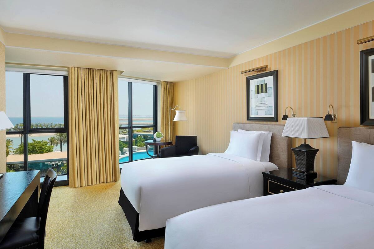 A hotel room with two beds, a desk, a chair, and a large window offering a view.
