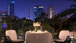An outdoor dining area with a table set for two, surrounded by wicker chairs, with a city skyline in the background.