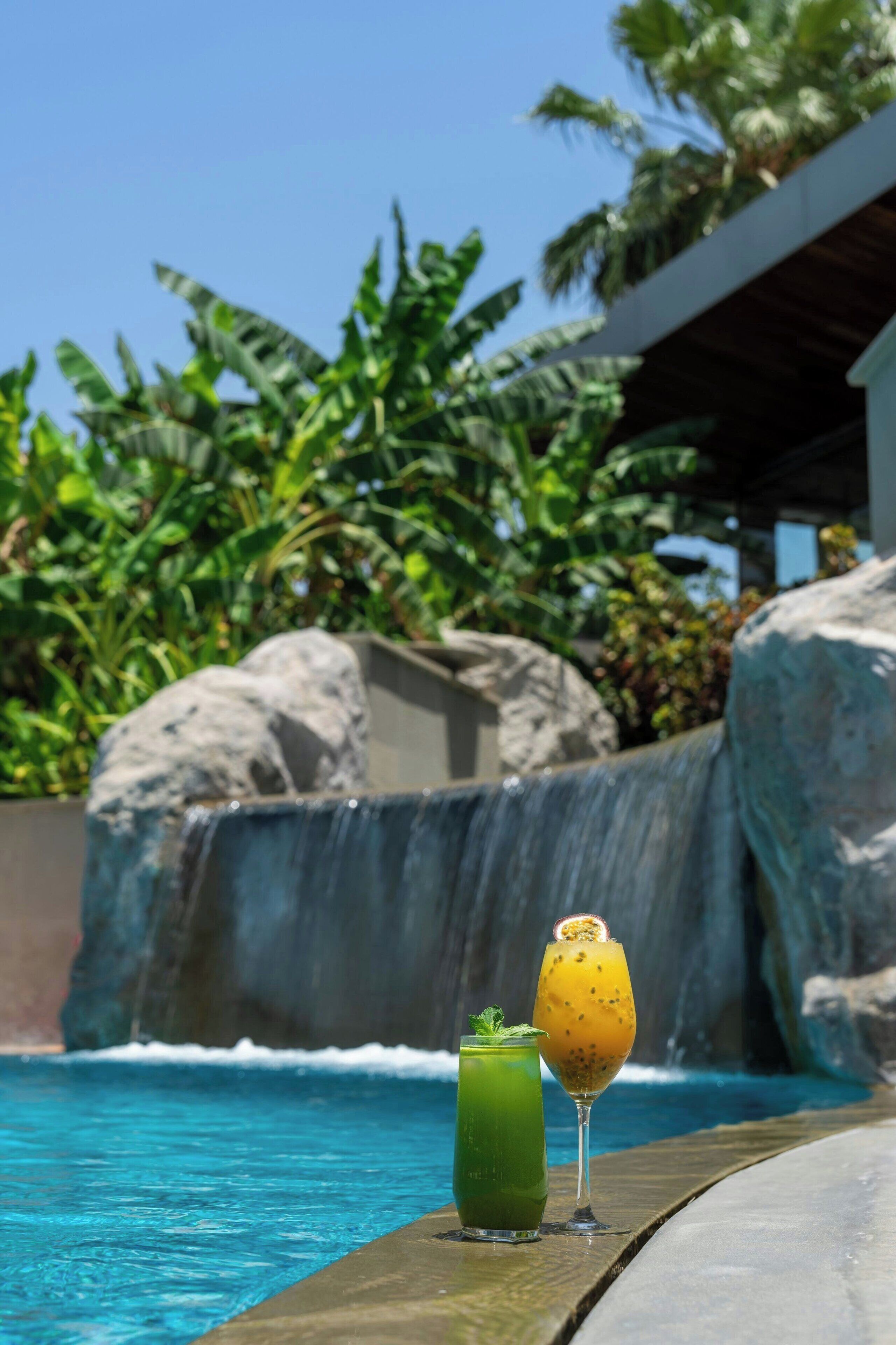 A poolside area with a clear view of the ocean, a waterfall, and lush greenery.