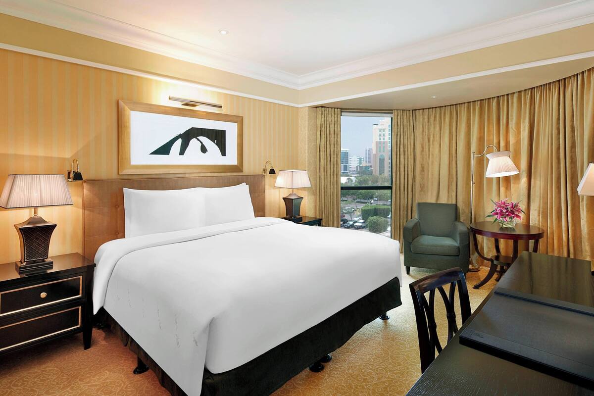 A hotel room with a large bed, two bedside tables with lamps, a desk with a chair, a view of the city through a large window, and a framed artwork on the wall.