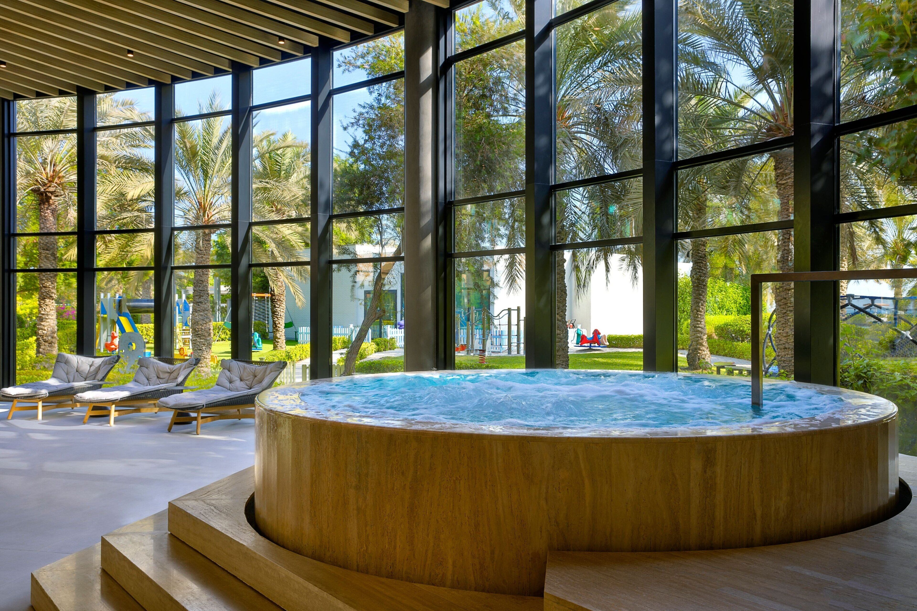 Un área de piscina interior moderna con jacuzzi, sillones de descanso y amplios ventanales con vistas a palmeras y un parque infantil.