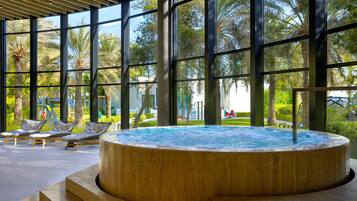 Un área de piscina interior moderna con jacuzzi, sillones de descanso y amplios ventanales con vistas a palmeras y un parque infantil.