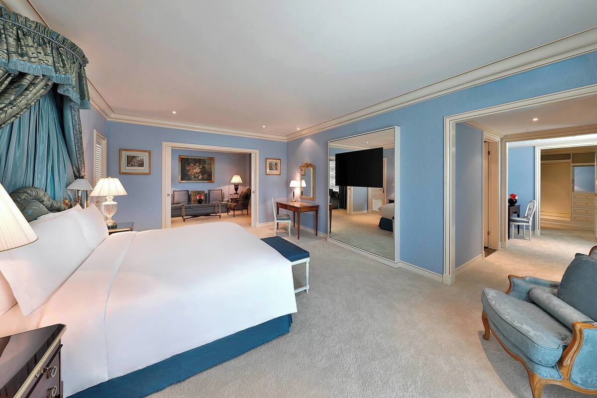 A hotel room with a large bed, a desk, a chair, and a sofa. There is a mirror on one wall and a door leading to another room.