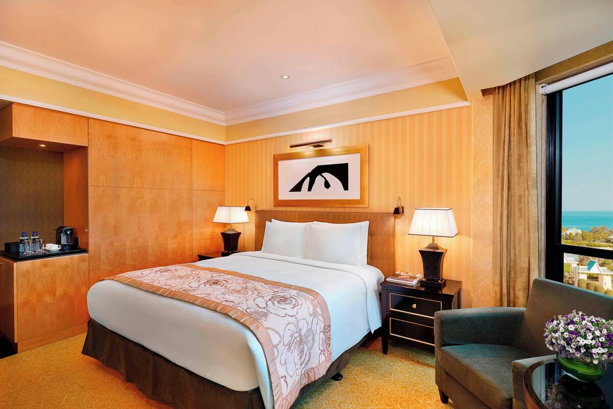 A hotel room with a large bed, two bedside tables with lamps, a chair, a view of the ocean, and a framed picture on the wall.