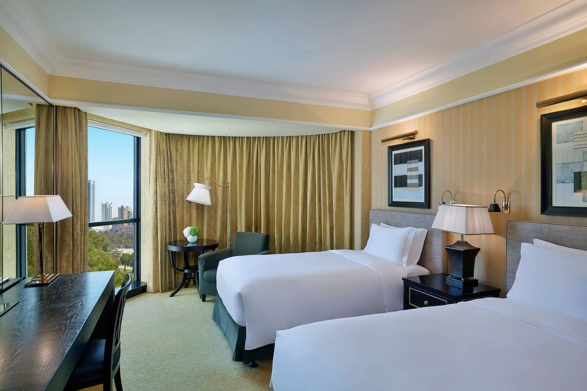 A hotel room with two beds, a desk, a chair, a small table, and a view of the city.