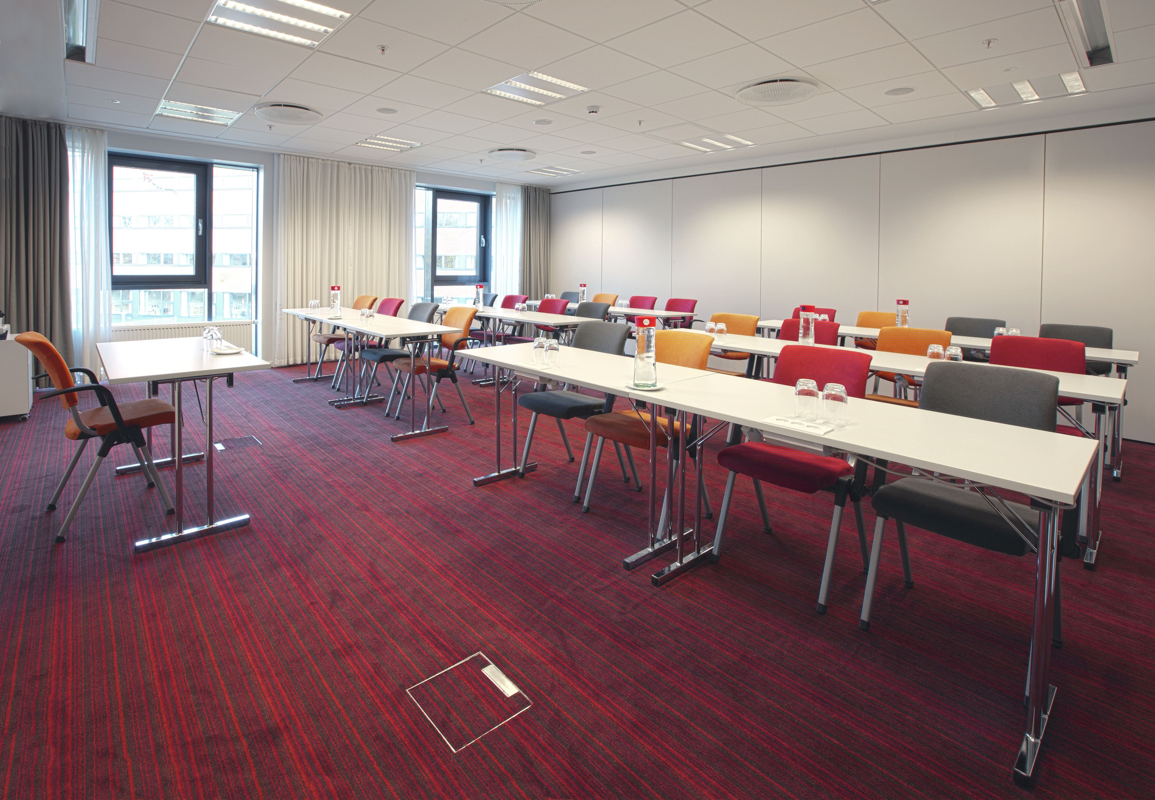 A conference room with multiple tables and chairs, carpeted floor, and large windows with curtains.