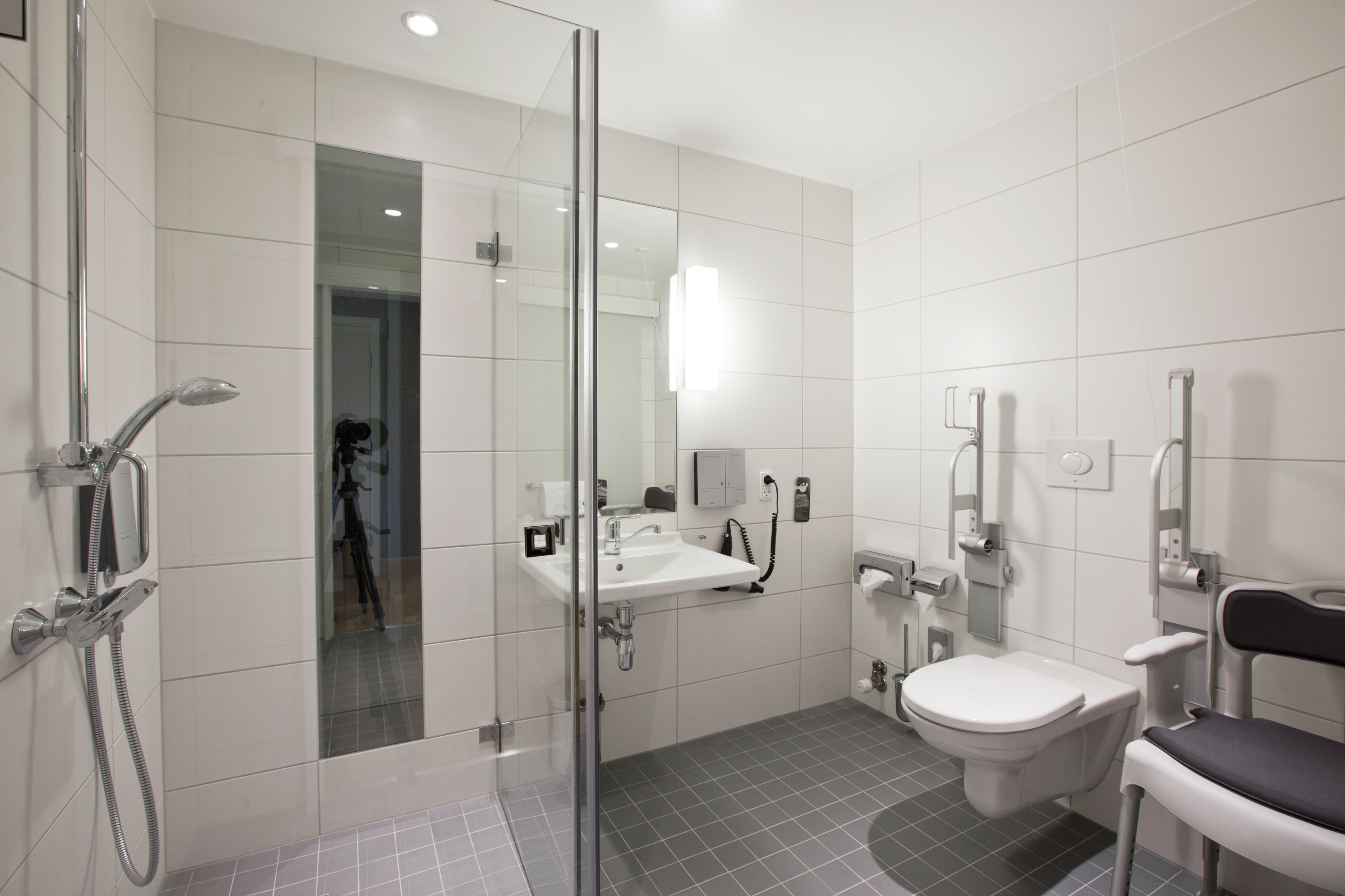 Un baño moderno con ducha envuelta en vidrio, un lavabo, un inodoro y una silla accesible para personas en silla de ruedas.