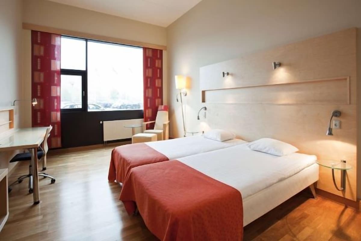 A hotel room with two beds, a desk, a chair, a window with curtains, and a lamp.