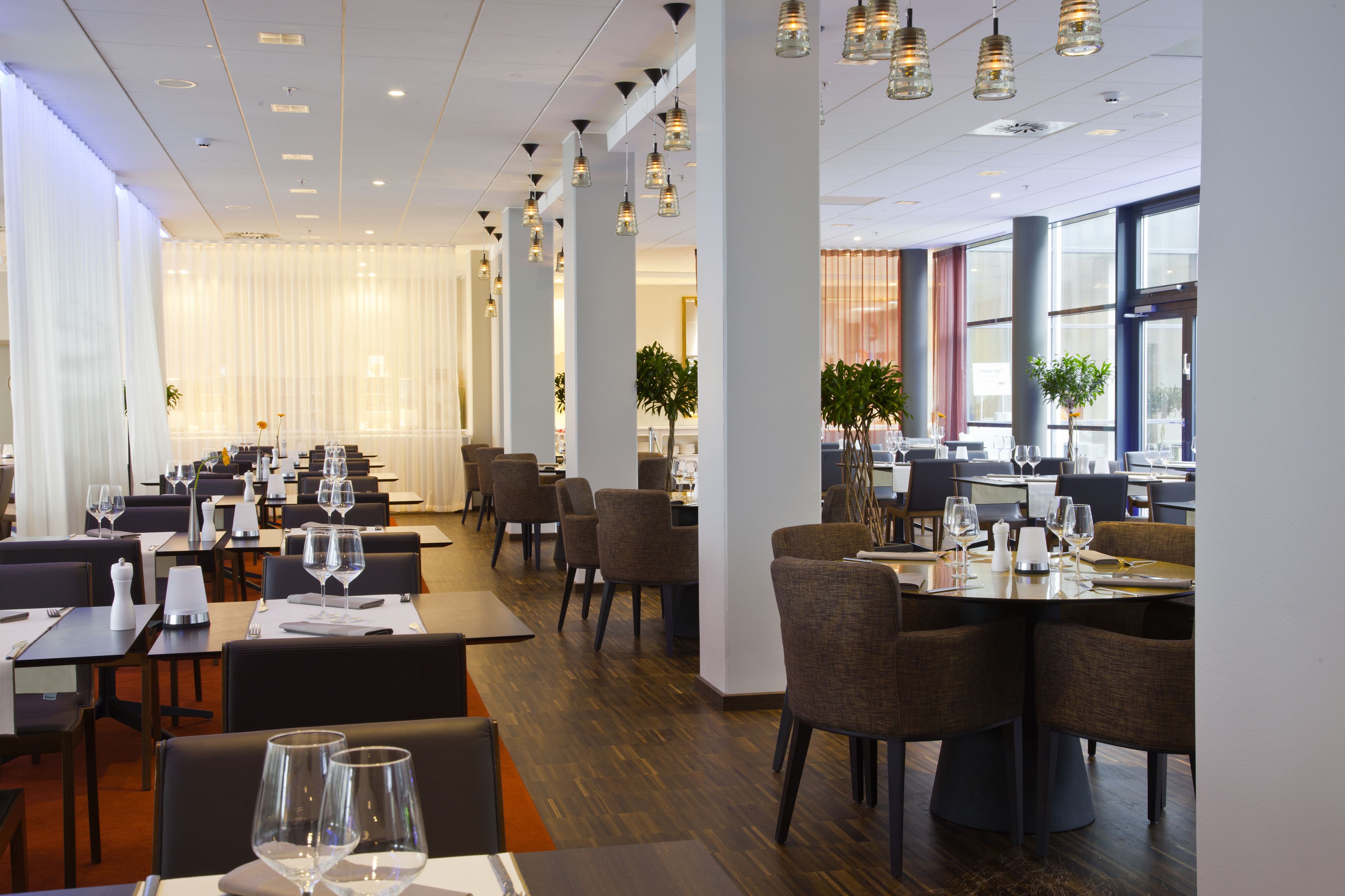 En modern restaurang med flera bord och stolar, stora fönster och hängande taklampor.