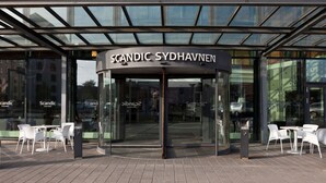 A modern building with a glass entrance and the name "Scandic Sydhavnen" above the doors. There are outdoor tables and chairs on the pavement in front. - Scandic Sydhavnen (Copenhagen)