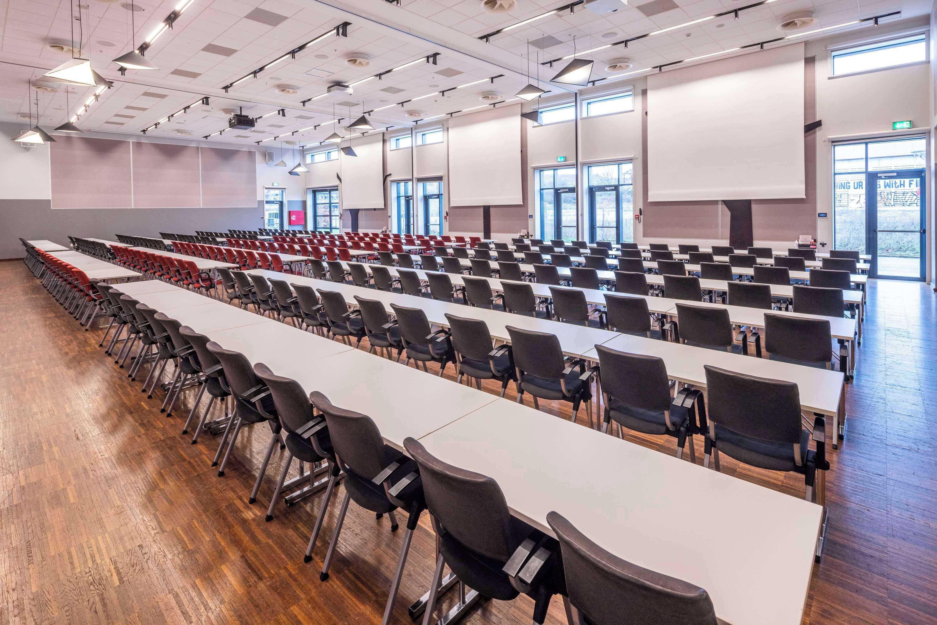 Un salón de conferencias con filas de mesas y sillas, ventanas amplias y una pantalla para proyector.