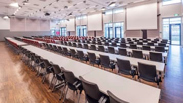 Un salón de conferencias con filas de mesas y sillas, ventanas amplias y una pantalla para proyector.