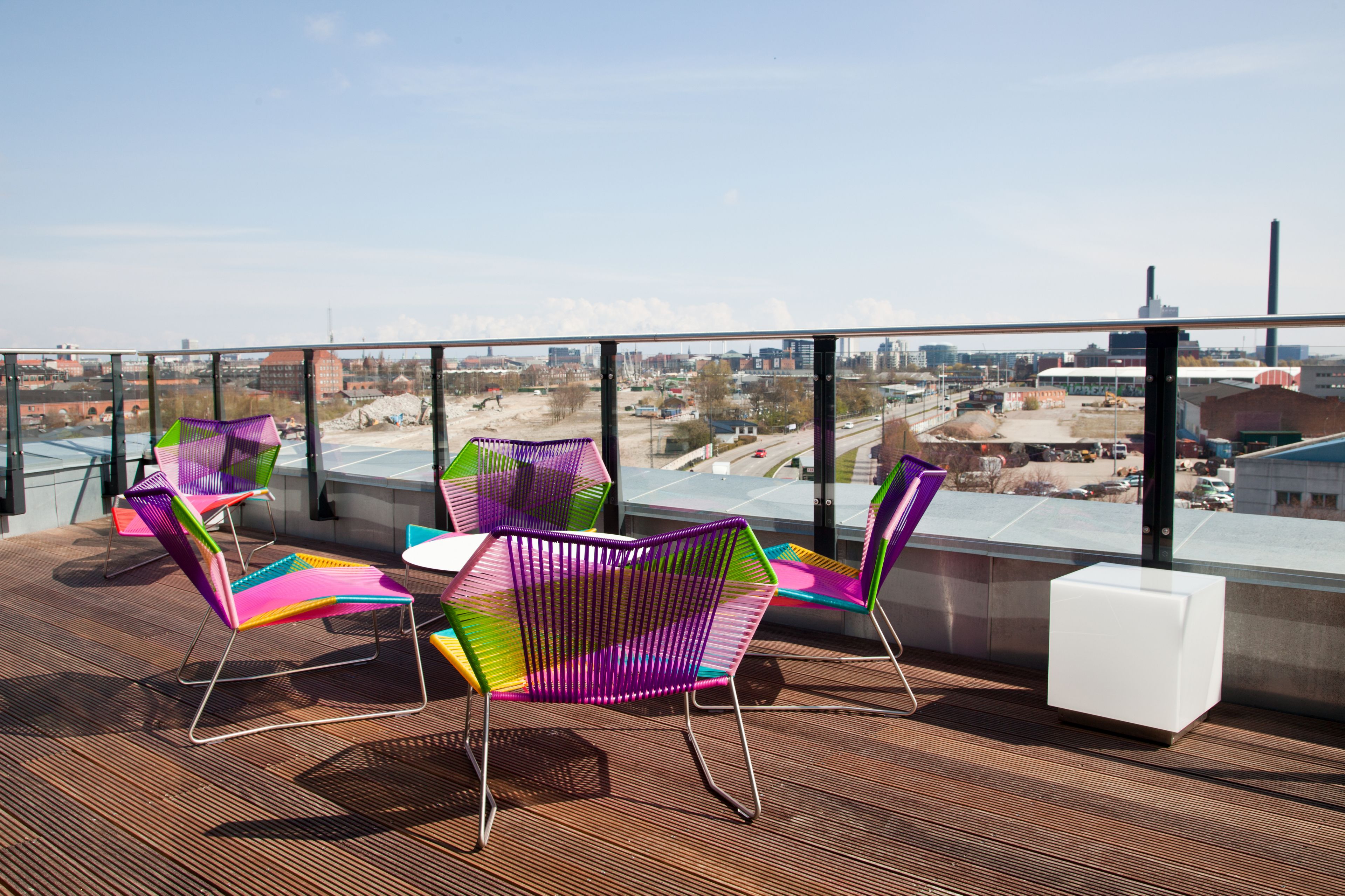 A rooftop terrace with colorful chairs and a white table, overlooking a cityscape.
