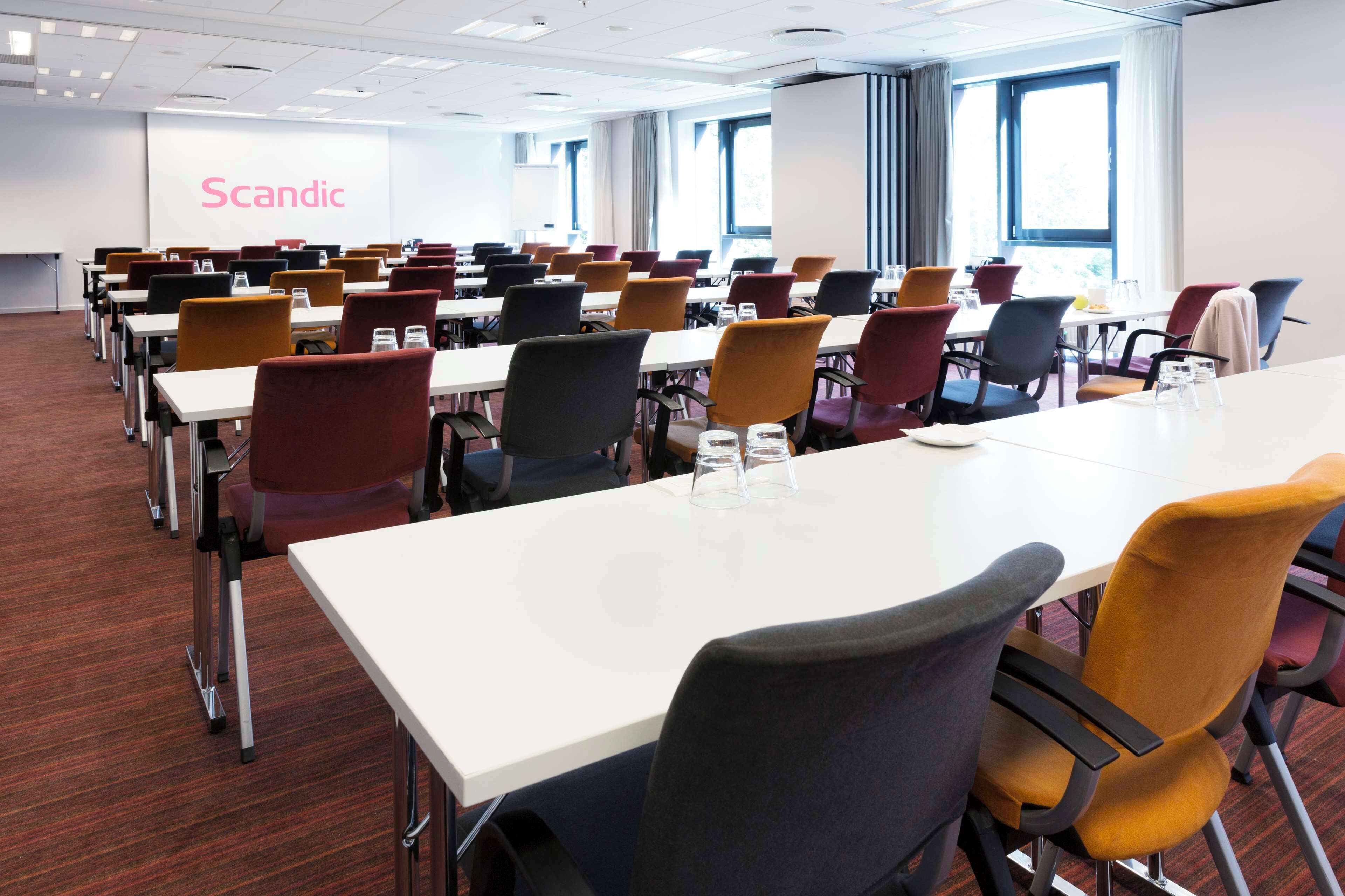A conference room with multiple tables and chairs, a projection screen with the word "Scandic," and large windows allowing natural light.