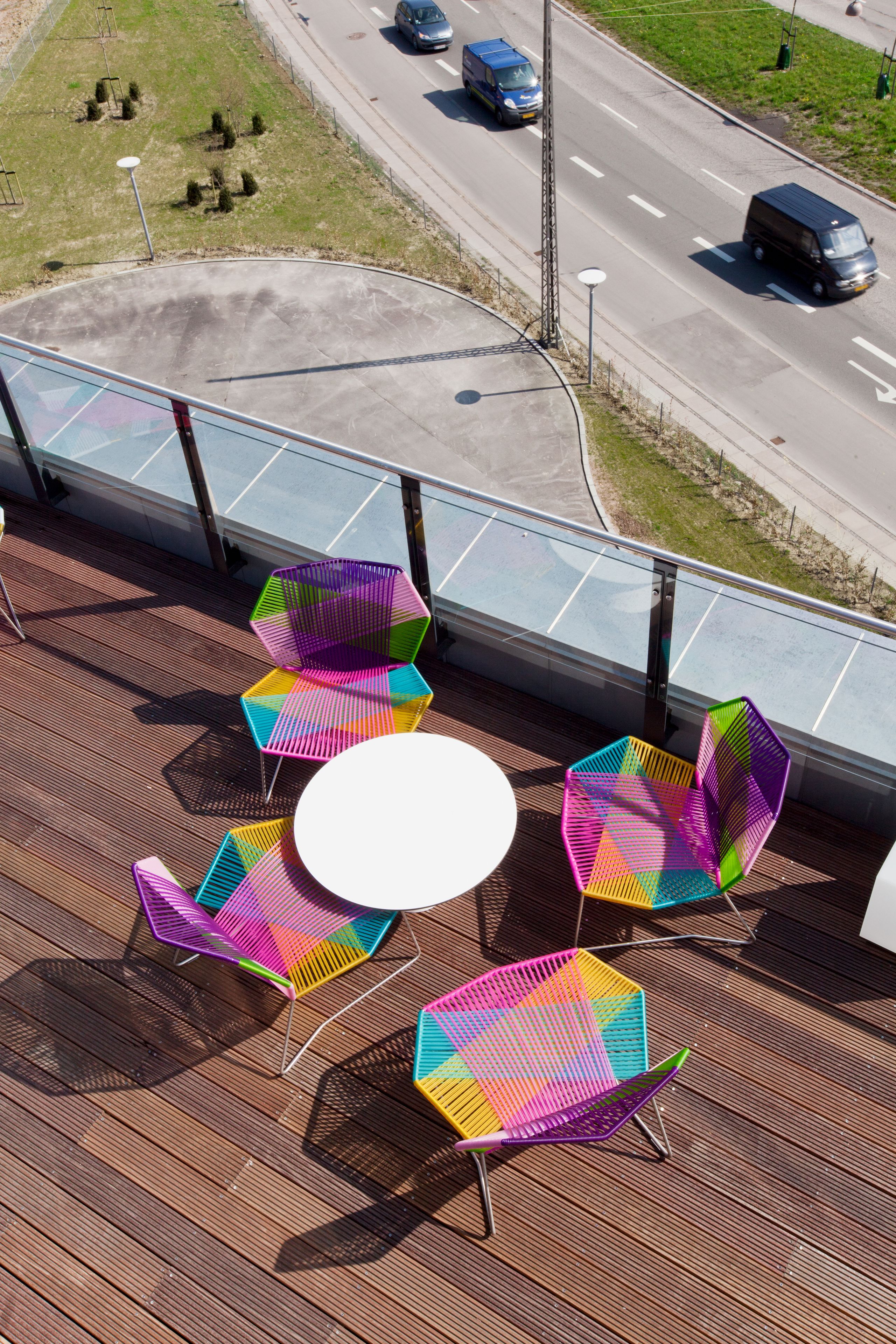En tagterrasse med farverige hexagonale stole og et rundt bord, med udsigt over en vej med køretøjer.