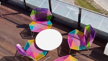 En tagterrasse med farverige hexagonale stole og et rundt bord, med udsigt over en vej med køretøjer.