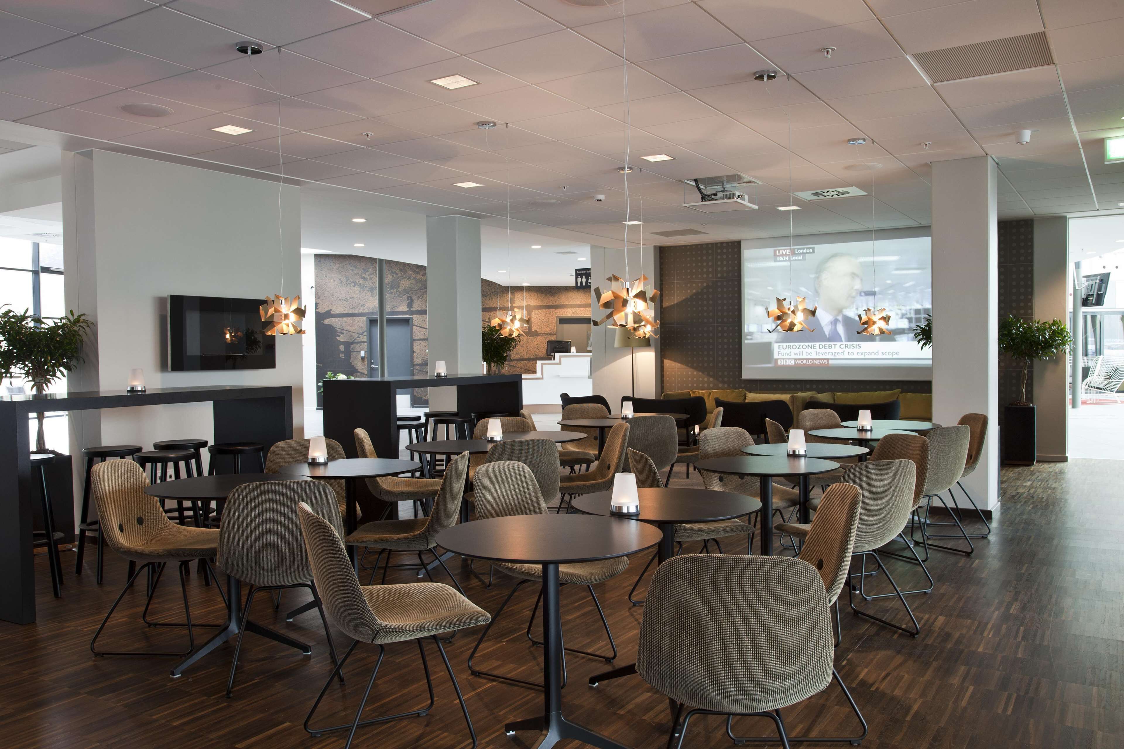 Un espace repas moderne avec des tables et des chaises rondes, un écran de télévision et un écran de projection diffusant les actualités.
