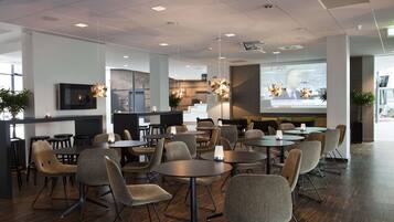 Un espace repas moderne avec des tables et des chaises rondes, un écran de télévision et un écran de projection diffusant les actualités.