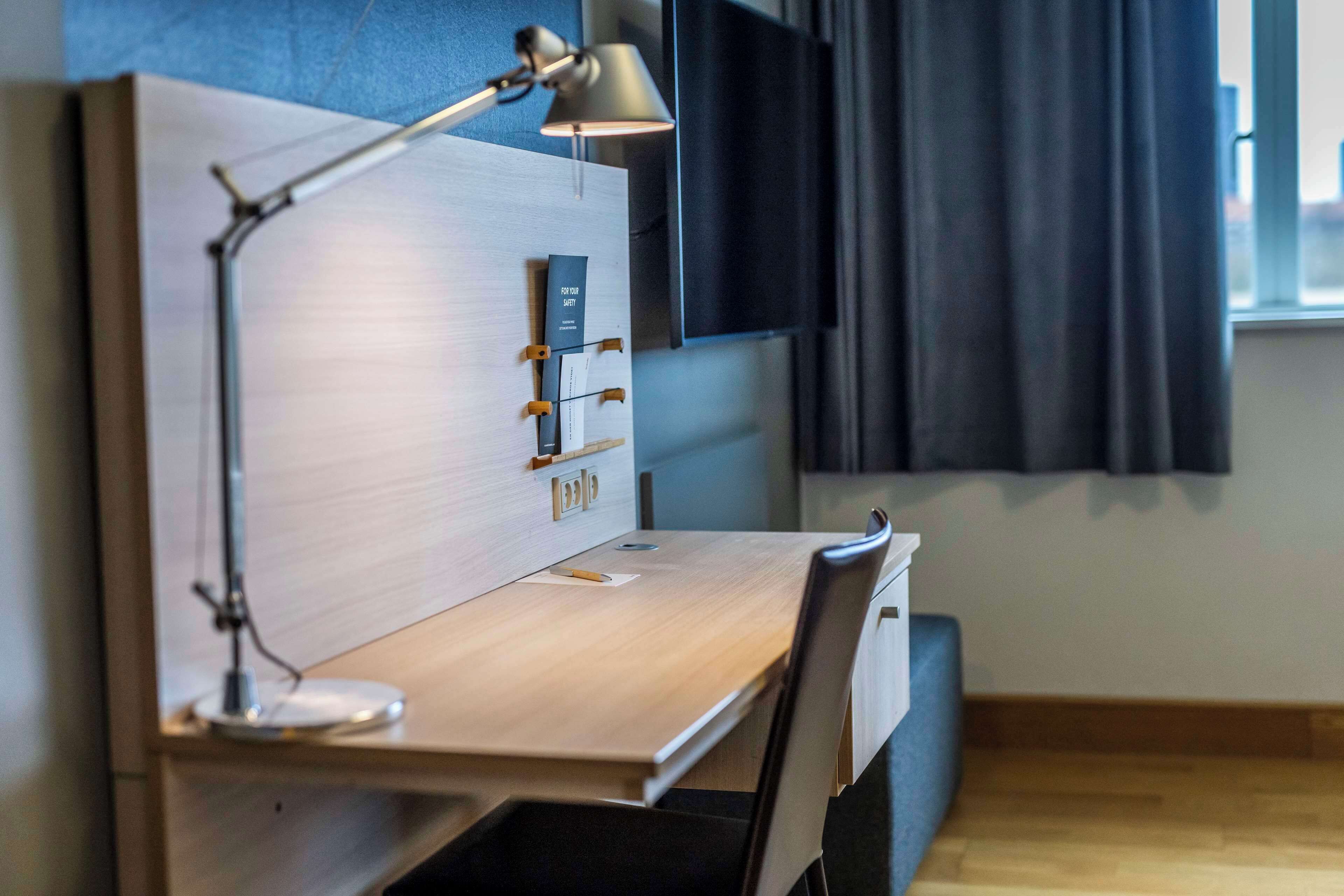 Ein moderner Arbeitsbereich mit Schreibtisch aus Holz, einer Lampe, einem Wand-TV und einem Fenster mit Vorhängen.